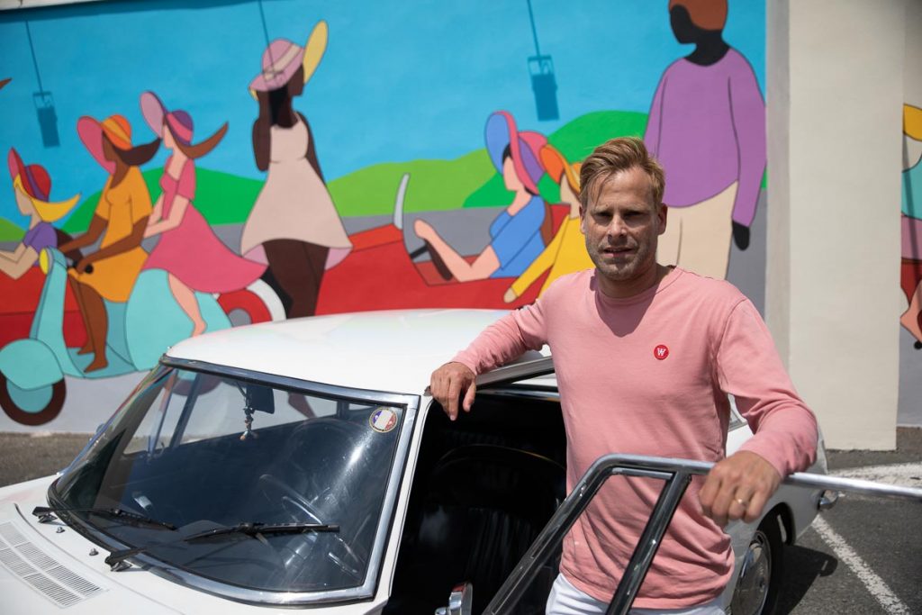 Franck Ayroles pose devant sa fresque murale et une Peugeot 404